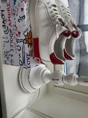 Dolce & Gabbana sneakers  - Snygga vita sneakers från Dolce & Gabbana med röda detaljer på häl och plös. Klassisk låg modell med vita snören och diskret logga. Tillverkade i läder med stilren design som passar perfekt till jeans eller shorts.