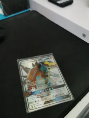 Turtonator GX Pokémonkort - Säljer ett Turtonator GX Pokémonkort med holografisk yta. Kortet har färgstark design med Turtonator i fokus och flera attacker som Shell Trap, Bright Flame och Nitro Tank GX. Kortet är inplastat i ett skyddsfodral och har klassisk Pokémon-baksida.