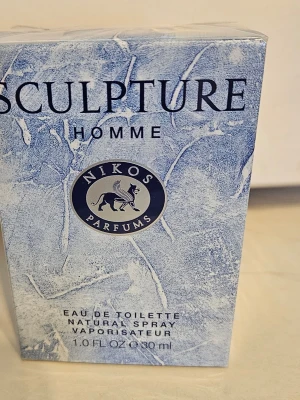 Nikos Sculpture Homme Eau de Toilette - Nikos Sculpture Homme Eau de Toilette är en parfym för män i en stilren blå och vit kartong med marmorliknande mönster. Flaskan innehåller 30 ml och har en naturlig sprayfunktion. Märket har en elegant logga med en bevingad lejonfigur.