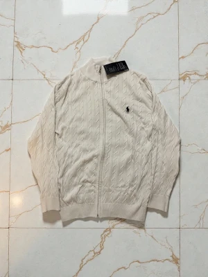 Vit kabelstickad ziptröja Polo Ralph Lauren - Kabelstickad vit ziptröja från Polo Ralph Lauren med hög krage och ribbade muddar. Tröjan har dragkedja framtill och den klassiska broderade loggan på bröstet. Perfekt för lager på lager och en clean look.