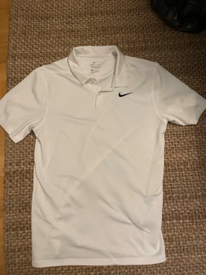 Vit piké från Nike i sportmarial  - Vit piké från Nike med klassisk krage och korta ärmar. Pikén har knappar framtill och en svart Nike-logga på bröstet. Tillverkad i ett lätt och sportigt sportmaterial som andas. Fick den i julklapp aldrig använd helt ny hör av vid frågor 