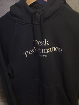Svart hoodie Peak Performance - Snygg svart hoodie från Peak Performance med broderad logga och text på bröstet. Klassisk känguruficka framtill och justerbar huva med snören. Perfekt för en avslappnad och sportig stil.