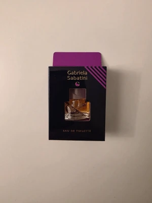 Gabriela Sabatini 3ml Eau de Toilette - En elegant Eau de Toilette från Gabriela Sabatini i en liten glasflaska med transparent lock. Doften kommer i en svart och lila förpackning med guldtext. Flaskan har en modern, vriden form och innehållet är ljust bärnstensfärgat.