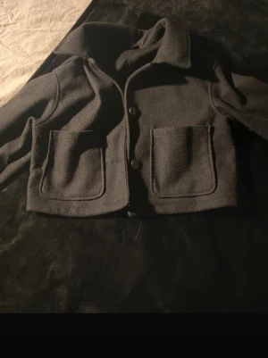 Svart cropped cardiganjacka - Snygg svart cardiganjacka med två stora framfickor och krage. Jackan är cropped och har en rak passform med knäppning framtill. Perfekt för lager-på-lager och enkel att styla till olika outfits.