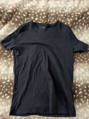 Svart basic t-shirt med rund hals - En enkel svart t-shirt med rund hals och korta ärmar. Perfekt för att matcha med jeans eller shorts. Materialet känns mjukt och lätt, troligen bomull. Passformen är normal och t-shirten har en klassisk look.