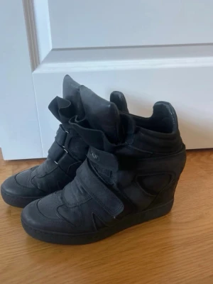 Svarta Carinii sneakers med kilklack - Säljer ett par svarta sneakers från Carinii med kilklack och högt skaft. Skorna har breda kardborreband och en unik, oversized plös i läder. Snygg streetstil som sticker ut och passar perfekt till jeans eller kjol.