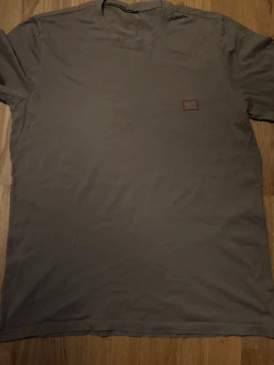 Grå boss t-shirt med liten patch - En enkel grå t-shirt i bomull med korta ärmar och en liten röd rektangulär patch på bröstet. Perfekt till vardags och lätt att matcha med andra plagg. Klassisk passform och rund halsringning.