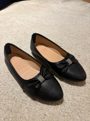 Svarta ballerinaskor med rosett - Snygga svarta ballerinaskor med spetsig tå och dekorativ rosett framtill, prydd med små glittrande stenar. Skorna har en slät yta i konstläder och ljus innersula. Perfekta för en stilren look. Passar mig som annars har storlek 39/40