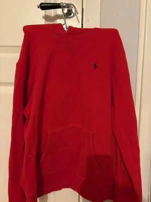 Röd hoodie från Polo Ralph Lauren - Röd hoodie från Polo Ralph Lauren med klassisk svart logga broderad på bröstet. Hoodien har huva och en stor magficka framtill. Tillverkad i mjukt bomullsmaterial och har ribbade muddar vid ärmslut och nederkant. 