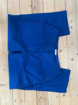 Blå byxor från Dedicated  - Säljer ett par blå byxor från Dedicated Brand med raka, hellånga ben och rena veck framtill. Modellen heter Pants Vallen Canvas Blue surf, är i slitstark ekologisk bomull och har en avslappnad passform. Sparsamt använda. Perfekta för en clean stil nu till våren och sommaren.