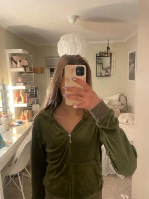 Olivgrön velour hoodie med dragkedja - Mjuk olivgrön hoodie i velourmaterial med dragkedja framtill och två fickor. Hoodien har en klassisk huva och ribbade muddar vid ärmslut och nederkant. Perfekt för en chill och avslappnad stil. Och är riktig