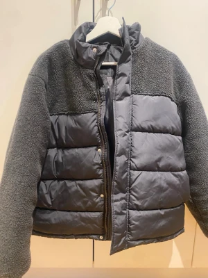 Grå pufferjacka från LMTD - Snygg grå pufferjacka från LMTD med quiltade partier och mjuka teddyärmar samt axelparti. Jackan har hög krage, dragkedja och tryckknappar framtill. Perfekt för kalla vinterdagar och ger en trendig look med sin mix av material. Priset går att diskutera 