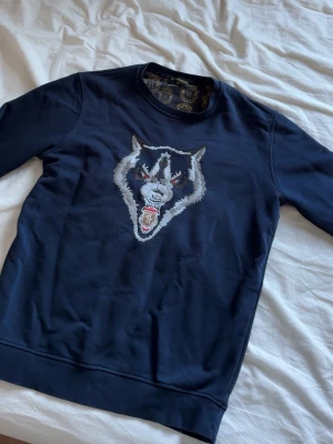 Mörkblå sweatshirt Billionaire M - Snygg mörkblå sweatshirt från Billionaire med ett stort broderat varghuvud på bröstet. Tröjan har rund halsringning och guldfärgade detaljer på insidan av kragen. Perfekt för dig som vill sticka ut med en cool design.