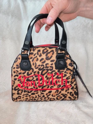 Leopardmönstrad Von Dutch handväska - Cool handväska från Von Dutch med leopardmönster i beige och brunt, svart konstläder på handtag och detaljer samt röd broderad logga framtill. Väskan har en kompakt, boxig form och dragkedja upptill. Perfekt statement piece för dig som gillar att sticka ut.