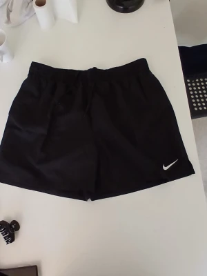 Svarta träningsshorts från Nike - Säljer ett par svarta träningsshorts från Nike med elastisk midja och klassisk vit logga på benet. Perfekta för gymmet eller löprundan. Lätta och bekväma med enkel design.