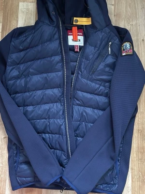 Mörkblå pufferjacka från Parajumpers - Snygg mörkblå pufferjacka från Parajumpers med huva och gul detalj vid halsen. Jackan har quiltad front, släta ärmar och bröstficka med dragkedja. Parajumpers-logga på vänster ärm. Perfekt för kalla dagar och riktigt stilren.