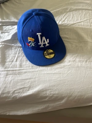 Blå LA Dodgers keps från New Era - Säljer en blå snapback keps från New Era, modell 59FIFTY, med LA Dodgers logga framtill och broderade detaljer i flera färger på sidan. Klassisk rak skärm och broderad New Era-logga på sidan. Perfekt för dig som gillar streetwear och sportig stil. Kostar 800kr ny p.g.a champions ”edition”. Pris ej hugget i sten.
