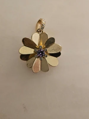 Blomformat halsband med lila sten - Unikt halsband med blomformat hänge i guldfärgad metall. Hänget har åtta blad och en lila sten i mitten, omgiven av små klara stenar. Perfekt för dig som gillar detaljerade och iögonfallande smycken.Smyckeshänge. använt ett par gånger.