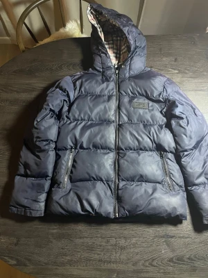 Mörkblå burberry pufferjacka med huva - Säljer en mörkblå pufferjacka från Burberry med klassiskt rutigt innerfoder i huvan. Jackan har två dragkedjeförsedda fickor framtill, stor huva och en patch med Burberry-logga på bröstet. Perfekt för kalla vinterdagar och riktigt snygg streetstil.