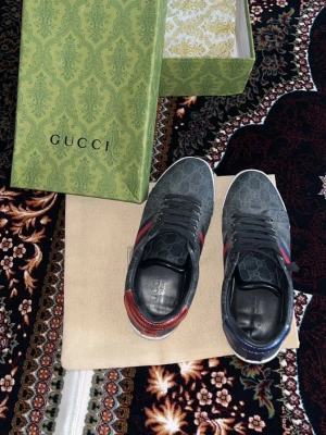 Gucci Sneakers Svartgrå - Skaffa dig ett par snygga Gucci sneakers i mörkgrått med klassiskt GG-monogram över hela skon. Skorna har blå och röda sidoremsor samt metallicdetaljer i rött och blått på hälen. Ovandelen är i canvas och insidan i svart läder. Perfekta för dig som vill sticka ut med lyxig streetstyle.