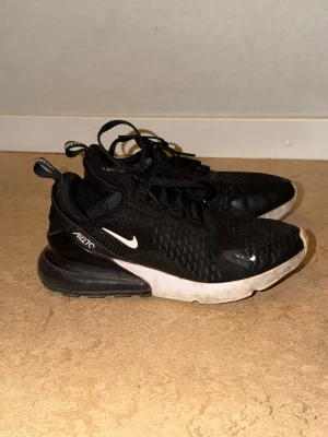 Nike Air Max 270 svarta sneakers - Svarta Nike Air Max 270 sneakers med vit sula och tydlig Air Max-dämpning i hälen. Ovandelen är i mesh för bra andningsförmåga och har vita detaljer som Swoosh-loggan och 270-tryck. Skorna har snörning och en sportig, modern look.