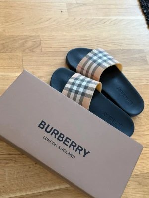 Burberry tofflor  - Pris kan diskuteras vid snabb affär.Snygga sandaler från Burberry med klassiskt beige, svart och rött rutigt band över foten och svart sula. Perfekta för sommaren och har den ikoniska Burberry-looken. Tillverkade i slitstarkt material och kommer med originalkartong.