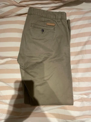 Beige chinos från Selected Home 34/32 - Snygga nästan helt nya beige chinos i storlek 34/32 från Selected Home med klassisk rak passform. Byxorna har bälteshällor, knappstängning och bakficka med knapp. Perfekta för en stilren och avslappnad look. Tillverkade i mjukt bomullstyg.