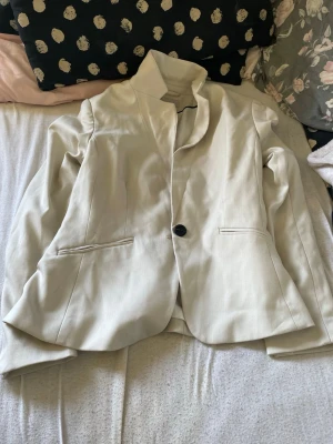 Beige kavaj med enkel knapp - Stilren beige kavaj med en knapp framtill och två fickor. Kavajen har en rak passform och klassisk krage. Materialet ser ut att vara en blandning av bomull och polyester med slät finish. Perfekt för en clean och modern look.