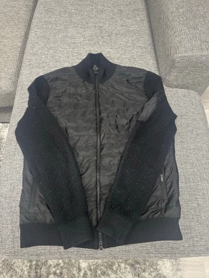 Svart Cardigan från J Lindeberg  - Säljer en svart pufferjacka med quiltad front och rygg samt stickade ärmar och muddar. Jackan har hög krage, dragkedja framtill och två sidofickor med dragkedja. Perfekt för kalla dagar och riktigt snygg till streetwear.