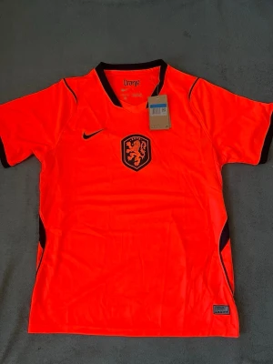 Holland Nike fotbollströja orange - Säljer en officiell Holland fotbollströja från Nike i starkt orange med svarta detaljer. Tröjan har KNVB-lejonet på bröstet, svart Nike-logga och texten 'Oranje' i nacken. Tillverkad i lätt och ventilerande material, perfekt för match eller träning.