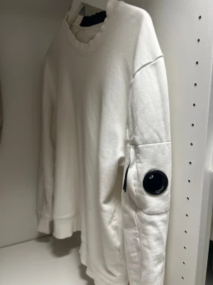 Vit sweatshirt från C.P. Company - Säljer en stilren vit sweatshirt från C.P. Company med rund halsringning och ribbade muddar. Tröjan har en ikonisk svart linsdetalj på ärmen samt en liten ficka med dragkedja. Perfekt för dig som gillar streetwear och enkel design.