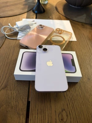 Lila iPhone 14 med tillbehör - Säljer en snygg lila iPhone 14 med två bakre kameror och stilren glasbaksida. Medföljer originalkartong, laddare och två skal – ett rosa och ett transparent. Perfekt för dig som vill ha en modern och fräsch mobil med cool design.