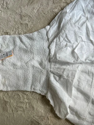 Vit axelbandslös sommarklänning - Säljer en söt vit axelbandslös klänning från Gina tricot med smock upptill och broderade blommor på kjolen. Perfekt för varma dagar och har en luftig, flowy passform. Klänningen är i bomull och har fina volangdetaljer längs kanten. Storlek 158/164 helt ny aldrig använd💖