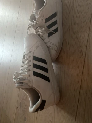 Vita Adidas sneakers med svarta ränder - Säljer ett par klassiska vita Adidas sneakers med de ikoniska tre svarta ränderna på sidan. Skorna har lågt skaft, vita snören och är tillverkade i syntetmaterial. Perfekta för en clean och sportig look.