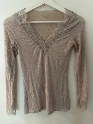 Beige långärmad topp med spets från Intimissimi - Supersnygg beige långärmad topp med v-ringning och spetsdetaljer vid halsringning och ärmslut. Tunn och mjuk, perfekt att styla med jeans eller kjol. Spetsen ger en extra touch av elegans och romantisk vibe. 💕💕