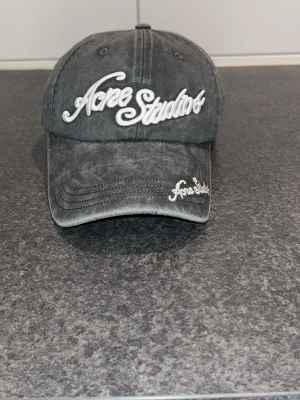 Grå keps från Acne Studios - Säljer en grå keps från Acne Studios med broderad logga i vitt framtill och på skärmen. Kepsen har en klassisk böjd skärm och justerbar baksida. Materialet är tvättad bomull som ger en avslappnad och trendig look.