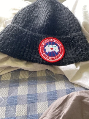  Canada Goose - Säljer en svart stickad mössa från Canada Goose med klassisk rund röd och vit logotyp framtill. Mössan är ribbstickad och har uppvikt kant för extra stilpoäng. Perfekt för kalla dagar och snygg till alla ytterkläder.