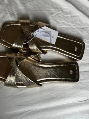 Guldiga sandaler / klackar med låg klack - Snygga guldiga sandaler från H&M med metallic finish och korsade remmar. De har en trendig fyrkantig tå och låg sula, perfekta för att lyfta vilken outfit som helst. Storlek 42 men passar 41