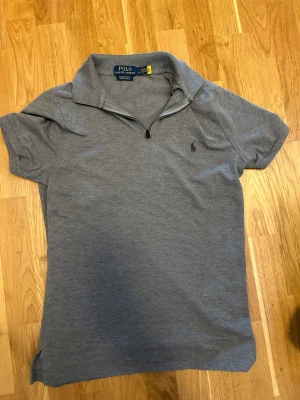 Grå polotopp från Polo Ralph Lauren - Snygg grå polotopp från Polo Ralph Lauren med korta ärmar och klassisk krage. Toppen har en liten dragkedja framtill och den ikoniska broderade loggan på bröstet. Tillverkad i mjuk bomull för en bekväm känsla.
