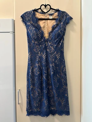 Blå spetsklänning med v-ringning - Snygg blå spetsklänning med nude underklänning och djup v-ringning. Klänningen har korta ärmar, figurnära passform och fina spetsdetaljer längs kanterna. Perfekt för dig som vill sticka ut med elegans och stil.