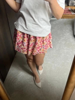 Unik blommig volangkjol i rosa och orange - Supersöt kort kjol med blommigt mönster i rosa, orange och vitt. Kjolen har volangdetaljer och en flowy passform som ger en lekfull vibe. Perfekt för varma dagar och enkel att styla med en basic t-shirt eller crop top. 