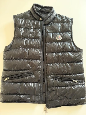 Svart dunväst från Moncler - Snygg svart dunväst från Moncler med glansig finish och ståkrage. Västen har två dragkedjeförsedda fickor framtill och klassisk Moncler-logga på bröstet. Tillverkad i lätt och värmande material, perfekt för lager-på-lager-stil. Ej några defekter, samt äkta. Strl: 1
