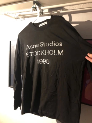 Svart långärmad t-shirt Acne Studios - Svart långärmad t-shirt från Acne Studios med tryck 'Acne Studios STOCKHOLM 1996' i vitt framtill. Klassisk rund hals och relaxed fit. Perfekt för dig som gillar stilrena och trendiga plagg från populära märken.