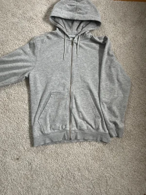 Grå zip hoodie från Weekday - Snygg gråmelerad hoodie från Weekday i storlek XS. Hoodien har dragkedja framtill, två stora fickor och justerbar huva med snören. Tillverkad i mjukt och bekvämt material, perfekt för en chill och avslappnad stil.