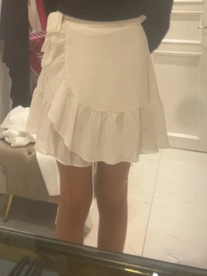 Vit omlottkjol med volang - Söt vit omlottkjol med volangkant och knytband i midjan. Kjolen har en kort och luftig modell med mjukt, tunt tyg som ger ett flowy intryck. Perfekt för en trendig och avslappnad look. Kjolen har också inbyggda shorts i.