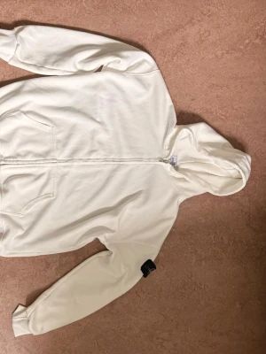 Vit hoodie från Stone Island - Säljer en clean vit hoodie från Stone Island med dragkedja framtill och klassisk huva. Hoodien har två fickor på magen och den ikoniska Stone Island-loggan på ärmen. Tillverkad i mjukt bomullsmaterial som känns skönt mot huden. Perfekt för dig som gillar stilrena plagg.