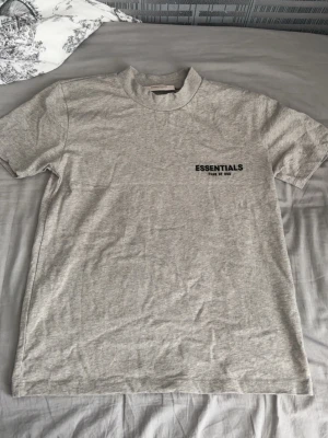 Grå Essentials Fear of God t-shirt - Snygg grå t-shirt från Essentials Fear of God med svart logga på bröstet och stor text på ryggen. Klassisk passform och rund halsringning. Perfekt till streetwear-stilen och enkel att matcha med allt. Tillverkad i mjuk bomull för skön känsla.