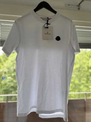 Vit Moncler t shirt med logga storlek 3 - Äkta vit t-shirt från Moncler med klassisk rund hals och svart logomärke på bröstet. Tillverkad i mjuk bomull för en skön känsla och perfekt passform. Enkel och stilren design som funkar till allt. Storlek 3