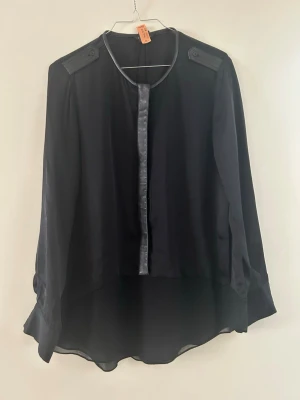 Club Monaco - lyxig svart blus - Svart blus med lång ärm och snygga axelknappar. Blusen har en rund halsringning och en dekorativa skinndetaljer. Materialet är tunt och lätt, 100% silke vilket ger ett flowigt och elegant intryck. Den är nyss kemtvättad därav sitter lappen kvar. 