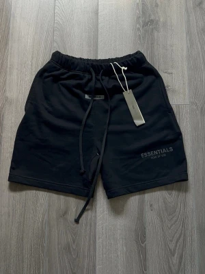 Svarta shorts från Essentials - Svarta shorts från Fear of God Essentials med elastisk midja och snörning. Mjuk bomullskänsla och diskret logga på vänster ben. Perfekta för en avslappnad och stilren look.